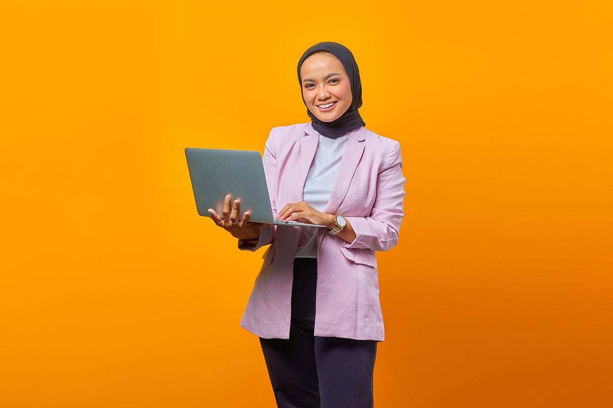 Portrait of smiling asian woman holding laptop and looking at camera over yellow background